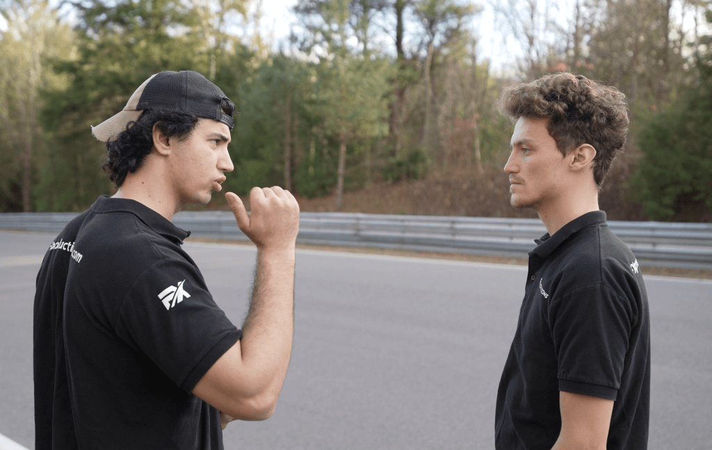 FX Productions Canada Two men in black shirts stand by a racetrack, one gesturing as the other listens, with trees and guardrail behind them—capturing a candid moment for FX Productions Canada, a trusted Toronto Video Production company. Toronto Video Production Company